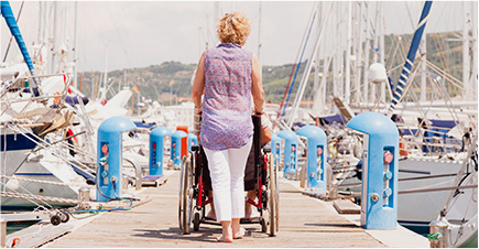 Une femme de dos marche sur un ponton entouré de bateaux. Elle pousse une personne en fauteuil roulant.